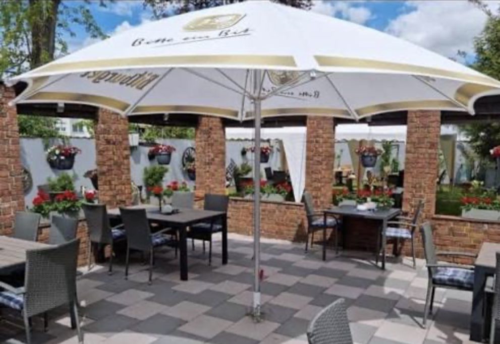 Gastronomischer Außenbereich mit Tischen, Stühlen und Sonnenschirmen, umgeben von Blumen.